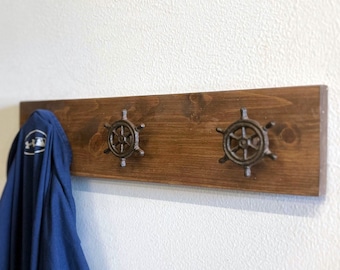 Könnte beinhalten: Ein Holz-Kleiderständer mit zwei Haken in Form von Schiffsrädern. Der Ständer ist dunkelbraun gebeizt und an einem der Haken hängt ein blaues Hemd.