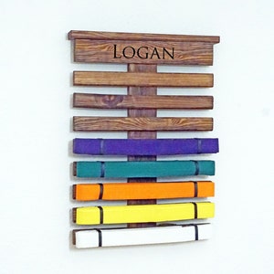 May include: A wooden belt rack with the name "LOGAN" engraved on the top. The rack has seven slots for belts, and there are six belts displayed in the colours purple, green, orange, yellow, and white.