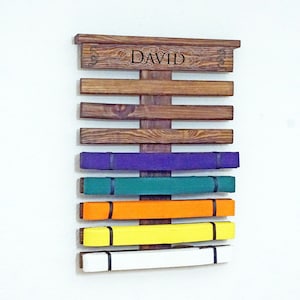 May include: A wooden belt rack with the name "DAVID" engraved on the top. The rack has 7 slots for belts and is holding 6 belts in various colours: purple, green, orange, yellow, and white.
