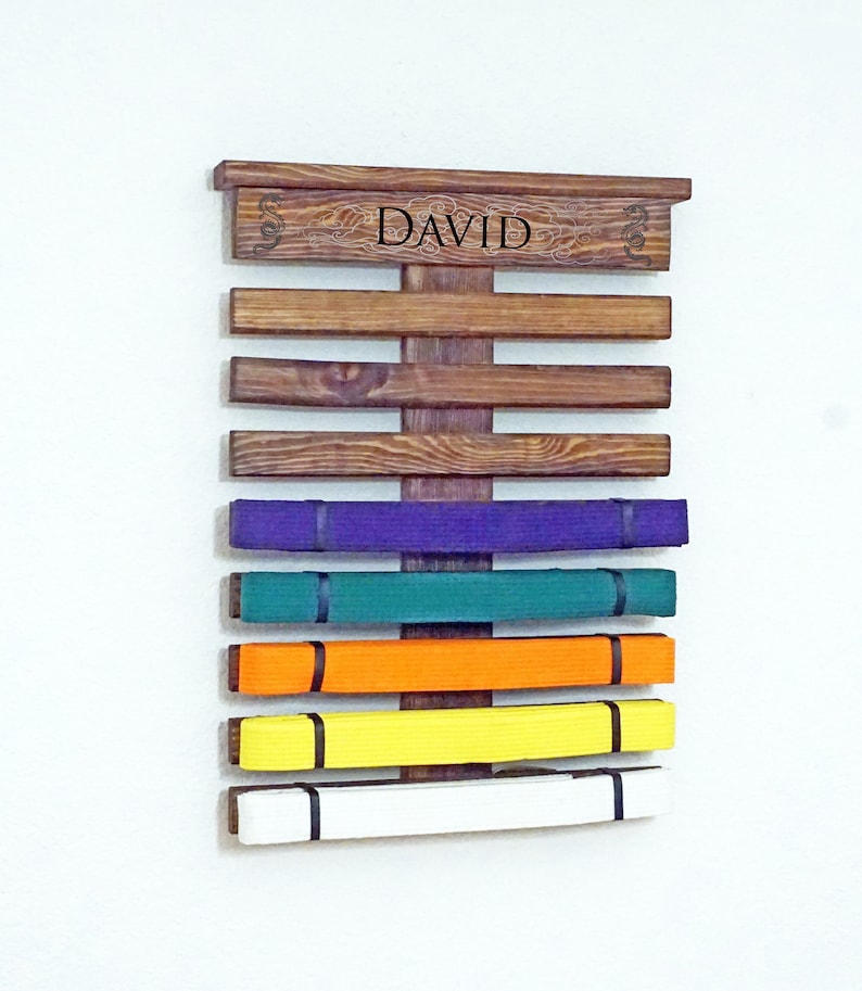 May include: A wooden belt rack with the name "DAVID" engraved on the top. The rack has 7 slots for belts and is holding 6 belts in various colours: purple, green, orange, yellow, and white.