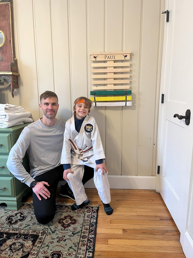May include: A father and son stand in front of a wooden karate belt rack with the name "PAUL" on it. The rack has several belts on it, including a green belt, a yellow belt, and a white belt. The son is wearing a white karate uniform with a yellow belt and an orange headband. The father is wearing a grey long-sleeved shirt and black trousers.