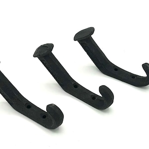 May include: Three black, rustic-style coat hooks. Each hook has a curved design with a rounded head and a curved hook end. The hooks are made of a dark, textured material, likely metal, and are mounted on a white background.
