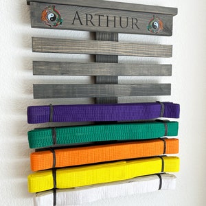 May include: A wooden martial arts belt display rack in a dark gray finish, with the name "ARTHUR" engraved on the top. The rack holds several colored belts: purple, green, orange, yellow, and white. Decorative emblems flank the name.