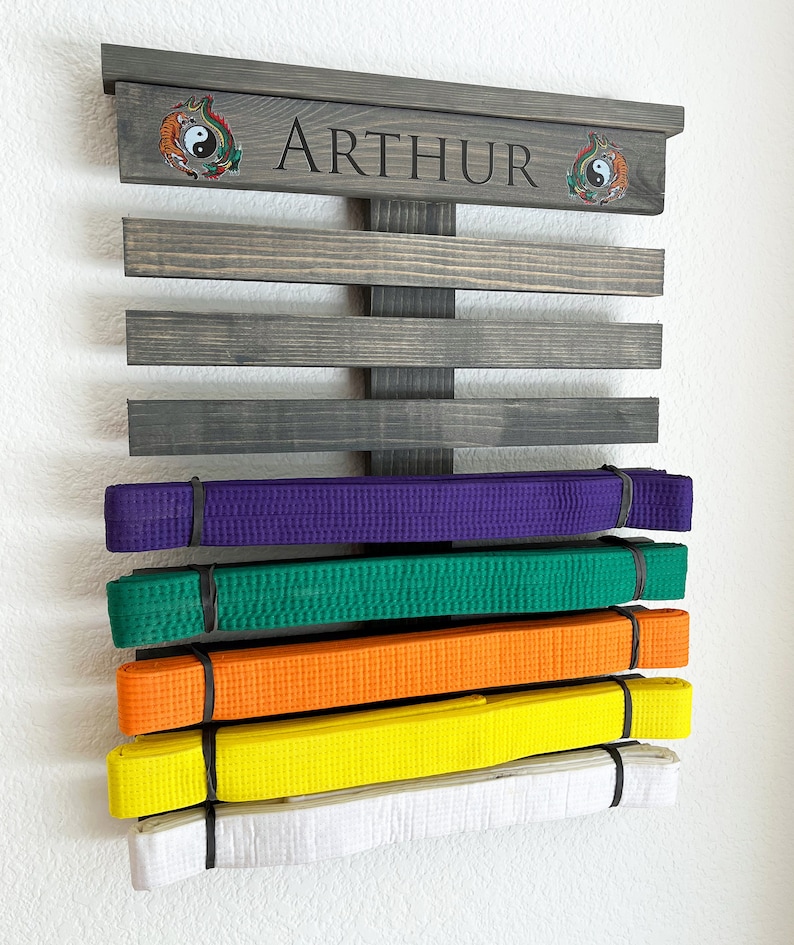 May include: A wooden martial arts belt display rack in a dark gray finish, with the name "ARTHUR" engraved on the top. The rack holds several colored belts: purple, green, orange, yellow, and white. Decorative emblems flank the name.