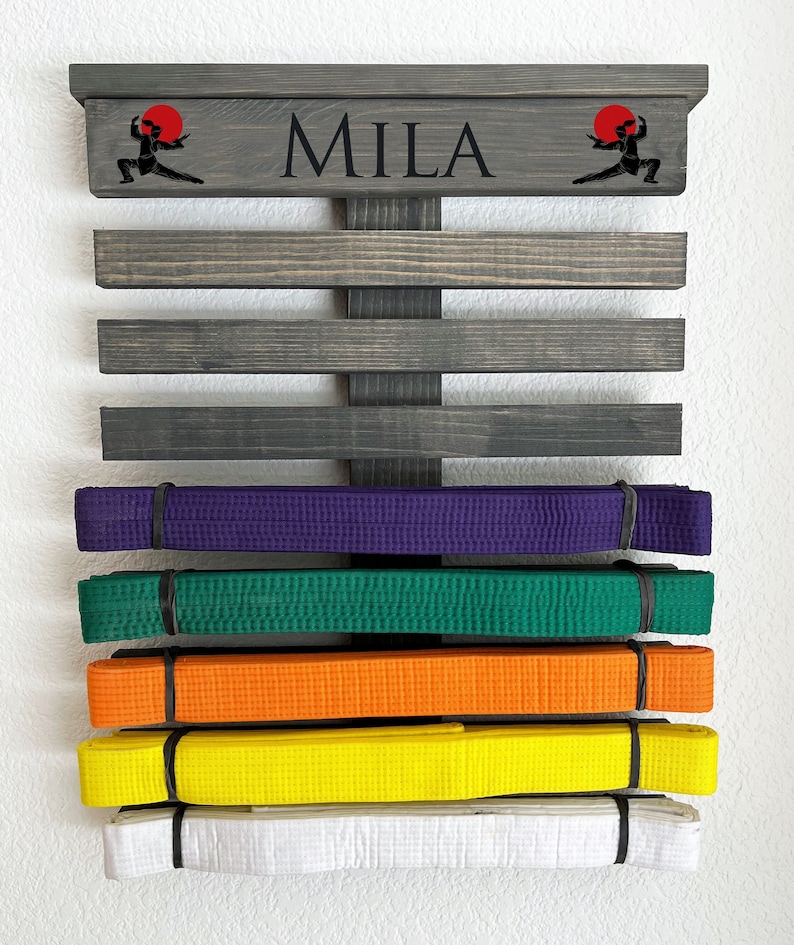 May include: A wooden martial arts belt display with the name "MILA" in black lettering. The display has several horizontal slats to hold belts of various colors, including purple, green, orange, yellow, and white. Ninja graphics are on the top.