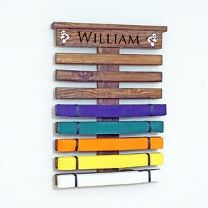 May include: A wooden belt rack with the name "William" on the top. The rack has 7 slots for belts and is holding a purple, green, orange, yellow, and white belt.