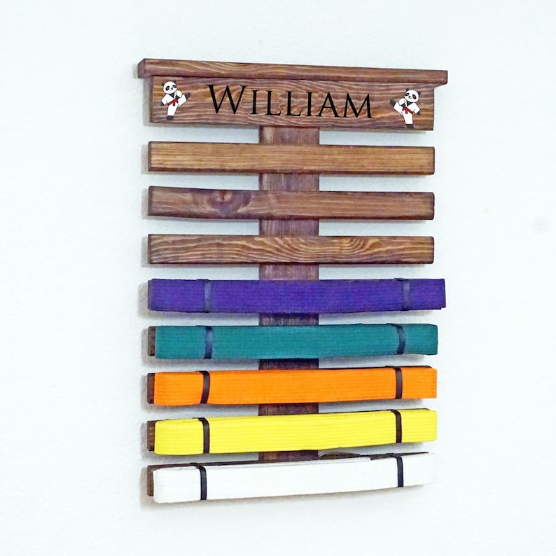 May include: A wooden belt rack with the name "William" on the top. The rack has 7 slots for belts and is holding a purple, green, orange, yellow, and white belt.