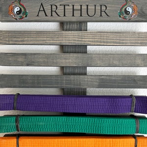 May include: A wooden belt display rack with the name "ARTHUR" in black lettering. The rack holds several colored martial arts belts, including purple, green, and orange. Decorative yin-yang tiger and dragon emblems are on the top.