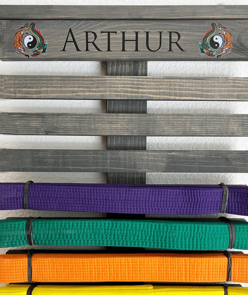 May include: A wooden belt display rack with the name "ARTHUR" in black lettering. The rack holds several colored martial arts belts, including purple, green, and orange. Decorative yin-yang tiger and dragon emblems are on the top.