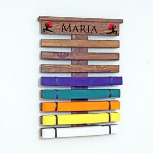 May include: A wooden martial arts belt rack with the name "MARIA" on the top. The rack has 7 slots for belts and is displaying a purple, green, orange, yellow, and white belt.