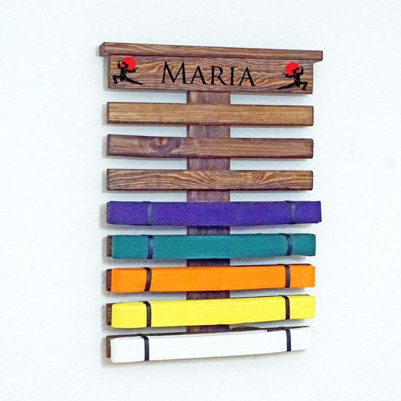May include: A wooden martial arts belt rack with the name "MARIA" on the top. The rack has 7 slots for belts and is displaying a purple, green, orange, yellow, and white belt.