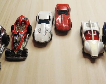 May include: A collection of six toy cars in various colors, including silver, red, white, and blue. The cars are all different styles, including a race car, a sports car, and a futuristic car.