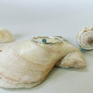 May include: A silver ring featuring a wavy band and a small, round, pale blue gemstone. The ring is presented on a large, cream-coloured seashell, with another shell in the background. The ring's design suggests an oceanic theme.