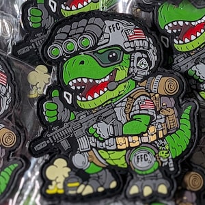 Puede incluir: Un parche táctico que muestra un dinosaurio de dibujos animados verde con equipo militar, incluyendo un casco con gafas de visión nocturna, un rifle y una mochila. El dinosaurio tiene la boca roja, dientes blancos y una bandera estadounidense.
