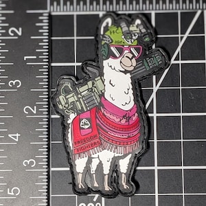 Può includere: Una toppa tattica raffigurante un lama bianco con casco verde, visore notturno, occhiali da sole rosa e un poncho rosso con la scritta FREEDOM FIGHTERS. La toppa è posizionata contro un righello che indica un'altezza di 7,6 cm.