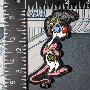 Op de afbeelding: Een roze cartoonmuis met een camouflagehelm en een groen-zwarte rugzak. De muis staat met gekruiste armen en heeft een rode neus. De tekst "FFC" staat op de staart van de muis.