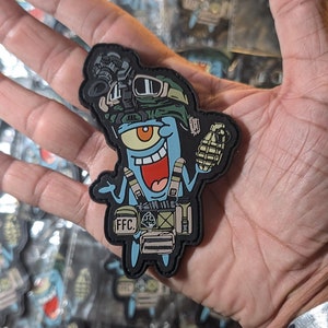 Puede incluir: Un parche bordado en negro y azul con un personaje de dibujos animados que lleva un casco militar, un chaleco y sostiene una granada. El personaje tiene un ojo grande y una amplia sonrisa. El parche dice "FFC" en el chaleco.