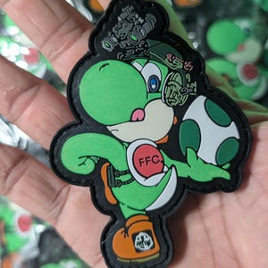 May include: A green Yoshi patch with a black border, wearing a military-style helmet and holding a green and white egg. The patch has the letters "FFC" on a red circle. The patch is approximately 3 inches tall.