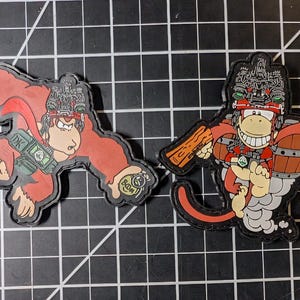 May include: Two embroidered patches featuring cartoon gorilla characters. One is red and tan, holding a bomb and a money bag. The other is red, tan, and white, with a barrel and wooden planks. Both have black borders.