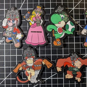 Op de afbeelding: Een verzameling kleurrijke geborduurde patches met personages uit een populaire videogamefranchise. De patches tonen personages als Mario, Princess Peach, Yoshi en Donkey Kong, met militaire ontwerpen. De patches liggen op een zwart rasteroppervlak.