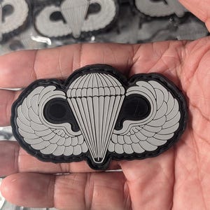 May include: A black and grey tactical patch featuring a central parachute design flanked by two stylized wings. The emblem is constructed with a raised, molded surface and a black border, showcasing a detailed canopy with vertical lines and symmetrical wing feathers.