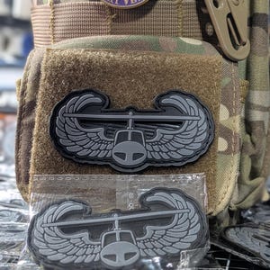 Puede incluir: Una bolsa táctica con un pin circular de Purple Heart Combat Veteran y dos parches de goma gris que representan un helicóptero con alas. Los parches tienen un acabado mate y se muestran tanto sujetos a la bolsa como dentro de plástico.