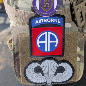 May include: A camouflage tactical pouch featuring three military patches: a circular Purple Heart Combat Veteran pin, a rectangular Airborne patch with a red background and white AA logo, and a black and grey paratrooper jump wings patch.