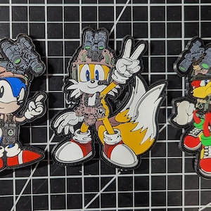 Könnte beinhalten: Drei gestickte Patches mit Charakteren aus dem Videospiel Sonic the Hedgehog. Die Patches sind im Militärstil gehalten, mit Tarnmustern und militärischer Ausrüstung. Die Charaktere sind Sonic, Tails und Knuckles.
