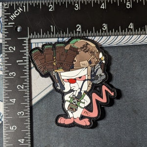 Puede incluir: Un ratón de dibujos animados negro y rosa con un casco militar con un patrón de camuflaje verde y marrón. El ratón sostiene un arma verde y marrón y tiene una cola rosa. El texto "FFC" está impreso en la pata del ratón.