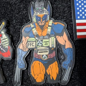 Op de afbeelding: Een blauw en oranje cartoonfiguur dat een tactisch vest draagt met de woorden "FFC", "STRENGTH & HONOR" en "FREEDOM FIGHTER" erop gedrukt. Het figuur draagt een helm met een groen vizier en heeft aan elke hand een paar klauwen.