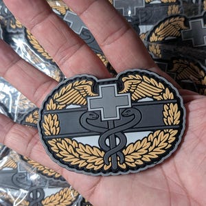 Puede incluir: Un parche táctico que presenta una cruz gris centrada sobre un símbolo de caduceo negro. El diseño está enmarcado por hojas de laurel de color beige y un borde gris. El emblema está fabricado en caucho moldeado con textura en relieve.