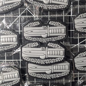 Puede incluir: Múltiples parches negros y grises con un cuchillo estilizado y un diseño rectangular, rodeados por una corona de laurel. Los parches están empaquetados en plástico transparente.