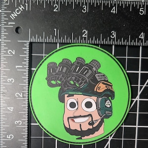 May include: A round, green sticker featuring a cartoon illustration of a person wearing a helmet with night vision goggles. The person has a beard and is wearing a headset. The text "FREEDOM FIGHTERS" is visible on the helmet.