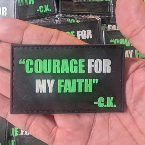 Puede incluir: Parche rectangular negro con el texto "COURAGE FOR MY FAITH" en blanco y verde. El parche también incluye las iniciales "-C.K." en blanco. El parche se sostiene en una mano.