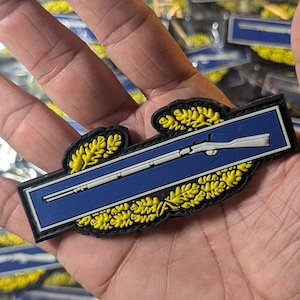 Peut inclure: Un écusson bleu et noir avec un fusil blanc. L'écusson est orné de feuilles de laurier jaunes et est rectangulaire avec des bords arrondis. L'écusson est tenu dans une main.