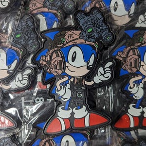 May include: A blue and tan embroidered patch featuring a cartoon character with blue eyes, a red nose, and a blue and white headband. The character is wearing a camouflage vest and holding a gun. The patch has the text "FFC" on it.