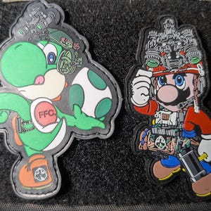 Puede incluir: Dos parches bordados con personajes de videojuegos. Un parche representa a Yoshi sosteniendo un huevo, y el otro muestra a Mario con equipo táctico. Ambos parches tienen diseños detallados y están delineados en negro.