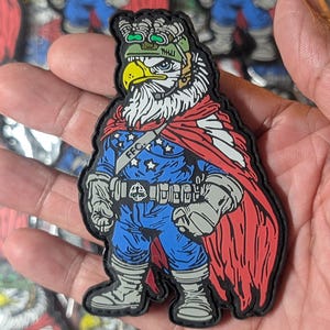 May include: A colorful patch featuring an eagle character in a superhero costume. The eagle wears a blue uniform with stars, a red cape, and a green helmet. The patch has the text "FFC."
