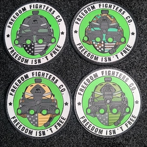 Może przedstawiać: Cztery okrągłe naszywki z zieloną obwódką i napisem "FREEDOM FIGHTERS CO." i "FREEDOM ISN'T FREE." Każda naszywka przedstawia wzór urządzenia noktowizyjnego z flagą amerykańską. Naszywki są na czarnym tle.