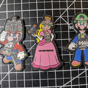 May include: Three colorful patches featuring characters from a video game franchise. Mario in red and blue, Princess Peach in a pink dress with the text "Freedom Fighters", and Luigi in blue and green. Each patch has a black border.