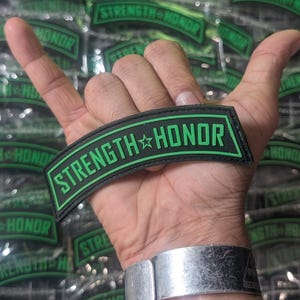 May include: A black and green patch with the text "STRENGTH HONOR" in green letters with a white star between the words. The patch is curved and has a stitched edge.