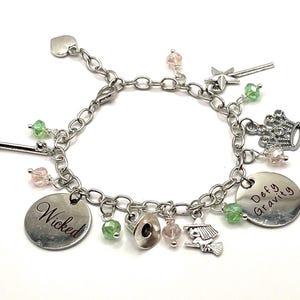 Puede incluir: Pulsera de abalorios plateada con abalorios que incluyen una bruja en una escoba, una corona, un sombrero y cuentas rosas y verdes. La pulsera tiene dos abalorios que dicen "Wicked" y "Defy Gravity".