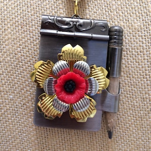 Pode incluir: Um colar de corrente dourada com um pingente de medalhão preto e dourado. O medalhão tem a forma de um pequeno livro com um desenho de flor vermelha na frente. Um pequeno lápis está preso ao medalhão.