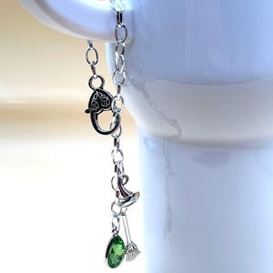May include: A silver-toned charm bracelet with a witch theme. The bracelet features a lobster clasp, a witch hat, a broom, and a green teardrop-shaped charm. The bracelet is hanging in front of a white mug.
