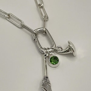 May include: Silver-toned necklace featuring a large oval clasp and a chain link design. The necklace has three charms: a broom, a green gemstone, and a horn. The charms are suspended from the clasp.