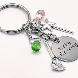 May include: A silver keychain with a charm that says "Defy Gravity". The charm is attached to a silver chain with a green glass bead, a pink glass bead, a silver star, and a silver broom.