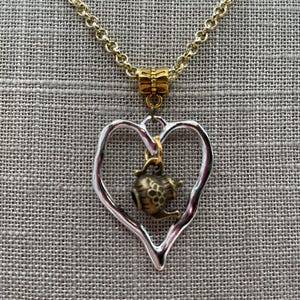 Può includere: Una collana a catena color oro con un ciondolo a forma di cuore irregolare color argento. Sospeso al centro del cuore c'è un piccolo ciondolo a forma di teiera color ottone antico. La collana include una contromaglia dorata per il ciondolo.