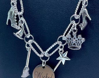 May include: Silver-toned necklace featuring charms related to the musical "Wicked." Charms include a high-heeled shoe, a crown, a star-shaped wand, a witch's hat, a broom, and a pendant engraved with the word "Wicked."