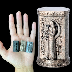 May include: Bronze-colored cylindrical statue of an Egyptian deity with hieroglyphs. Three miniature statues are held in a hand, each with a similar design. The larger statue features a detailed depiction of the deity with a bird head and crescent moon.