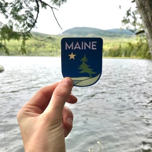 May include: A blue and white sticker with a pine tree and a star, and the word "MAINE" in white letters. The sticker is being held in front of a lake and mountains.